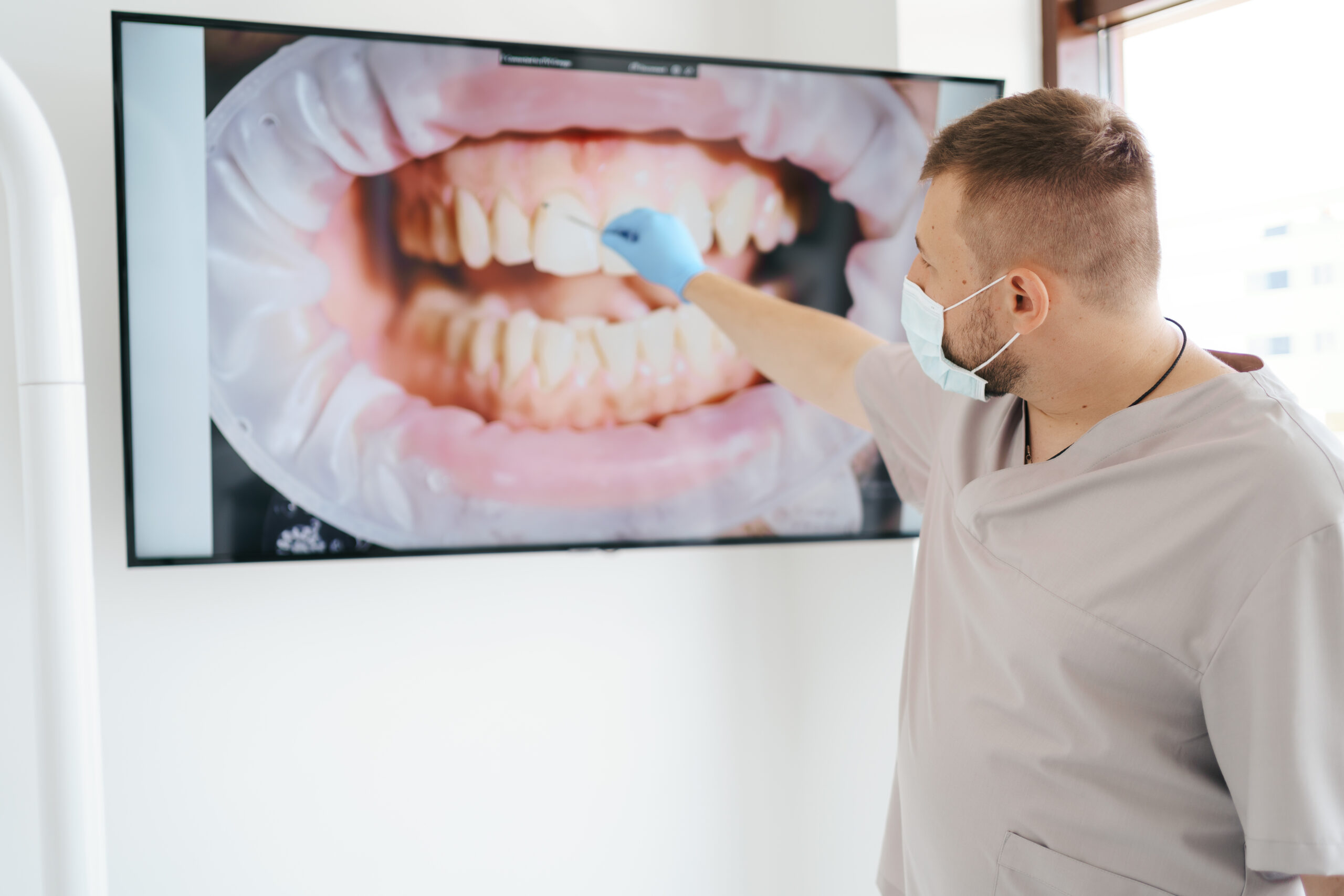Dentist in medical mask pointing on the patient teeth on a big screen explaining treatment phases